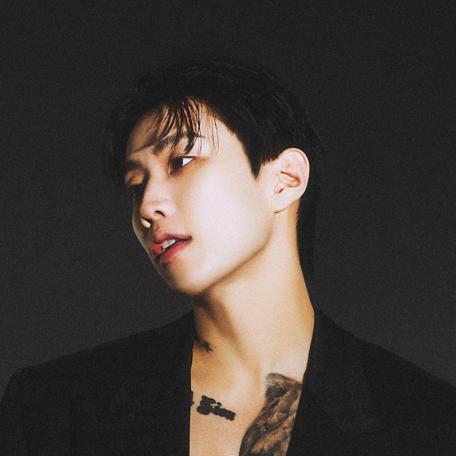 A person with short dark hair gazes to the side, wearing a black jacket over bare skin, revealing tattoos on their chest and collarbone. The background is dark and plain.