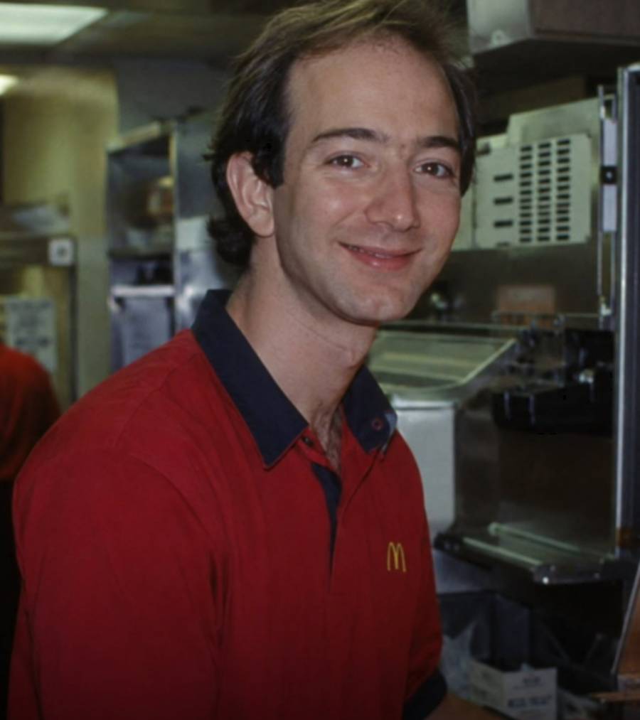A man wearing a red McDonald's uniform shirt smiles while standing in a kitchen area with stainless steel equipment and shelves behind him.