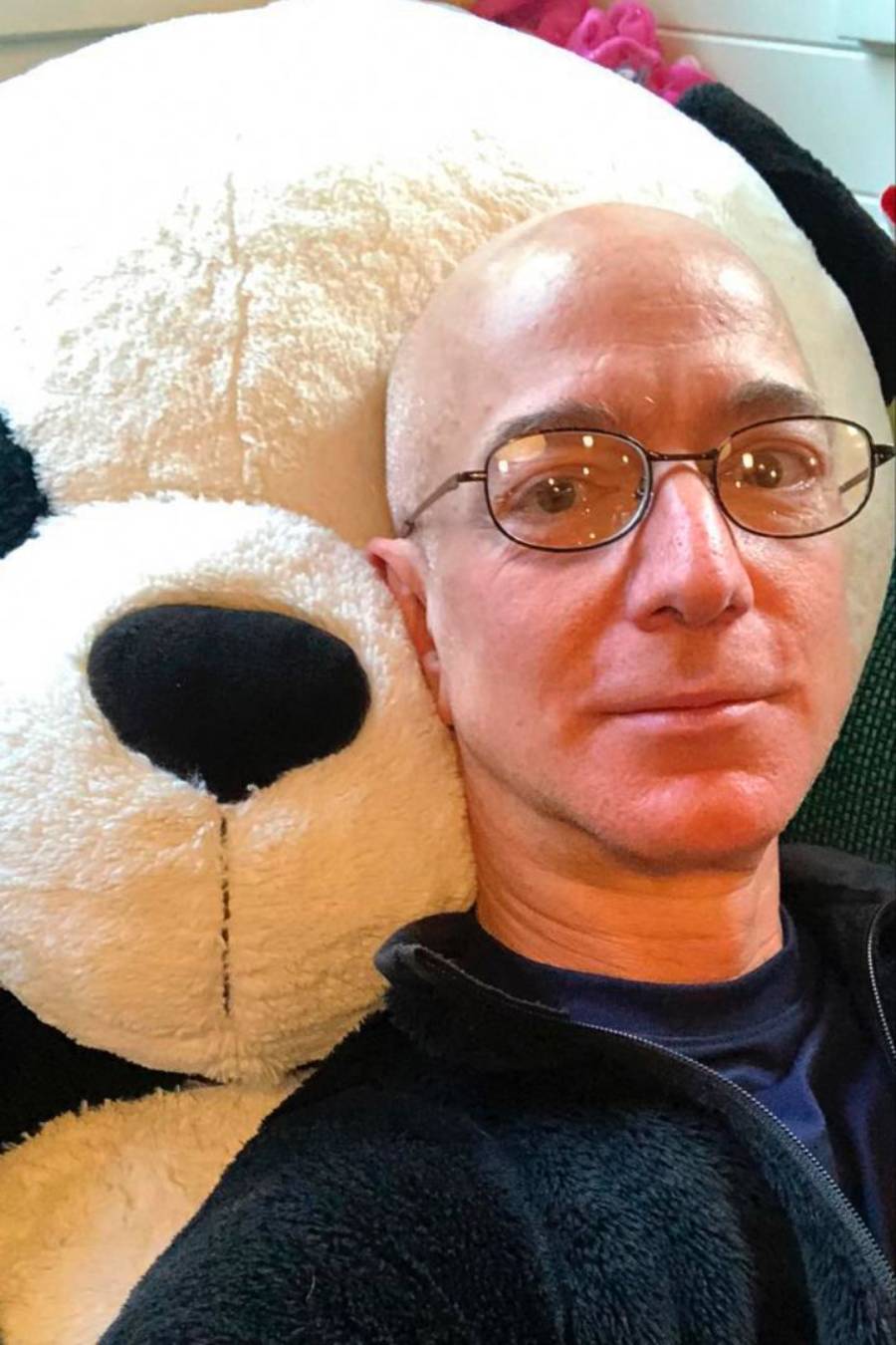 A man with glasses and a black jacket is taking a selfie while leaning against a large stuffed panda bear. The man is smiling slightly, and the panda's face is close to his.