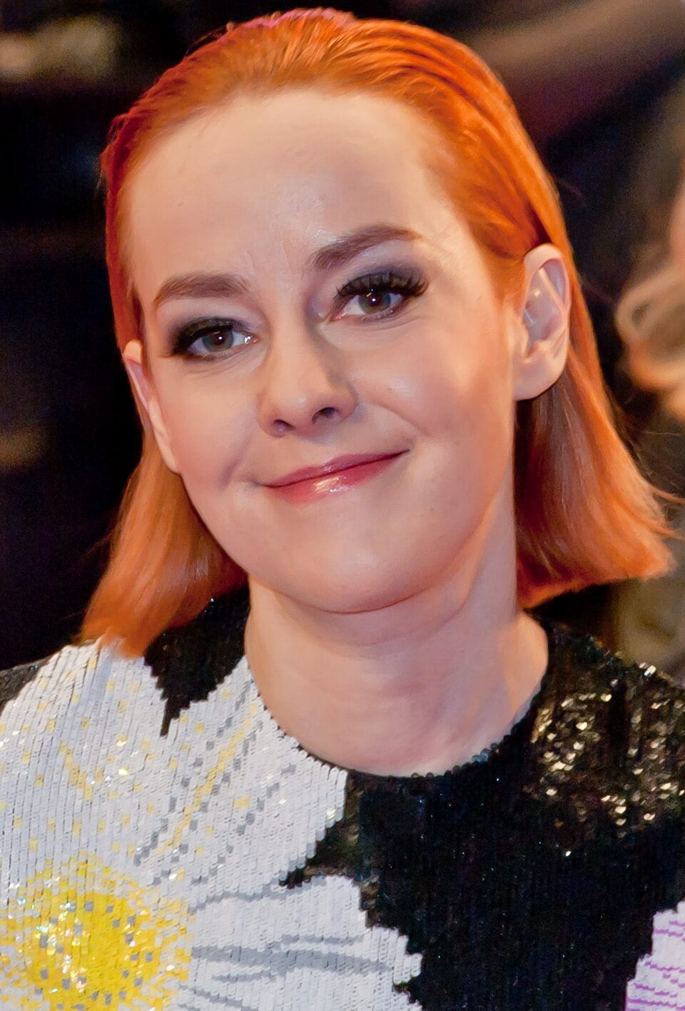 A woman with straight, shoulder-length red hair and fair skin is smiling softly. She is wearing a sequined outfit featuring a large white and yellow flower pattern. The background is dark and out of focus.