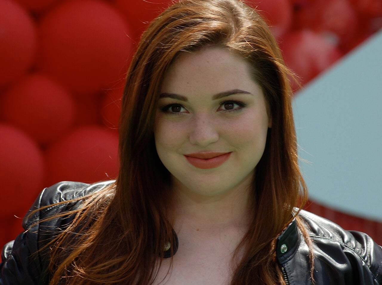 A young woman with long red hair and fair skin smiles softly at the camera. She is wearing a black leather jacket, and the background features a cluster of red balloons.