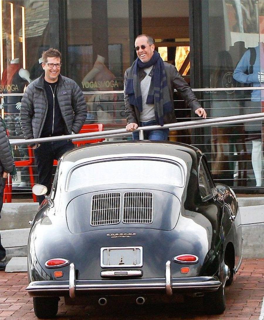 Two men stand smiling and chatting behind a vintage black Porsche parked on a brick street, outside a modern shop with glass windows and a sign that reads "YOGASMOGA.
