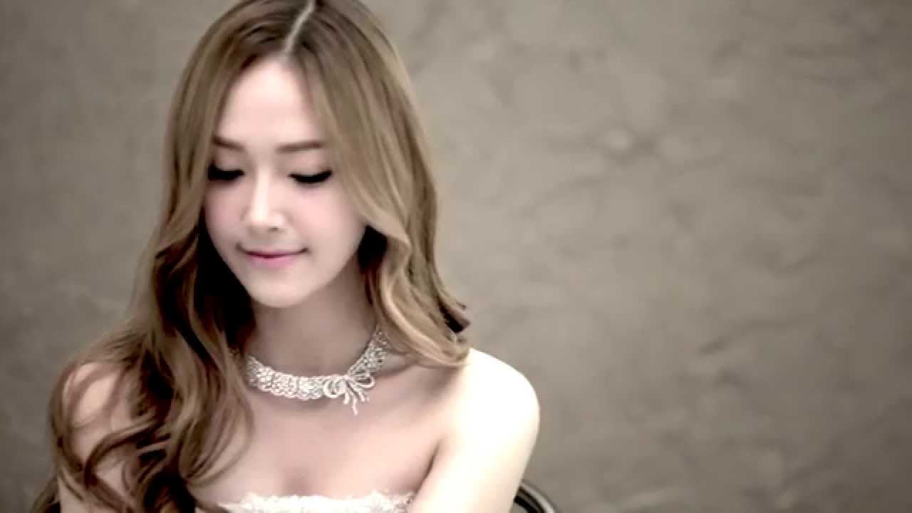 A woman with long, wavy light brown hair looks down and smiles softly. She wears a white lace dress and a delicate lace choker necklace. The background is a plain, beige textured wall.
