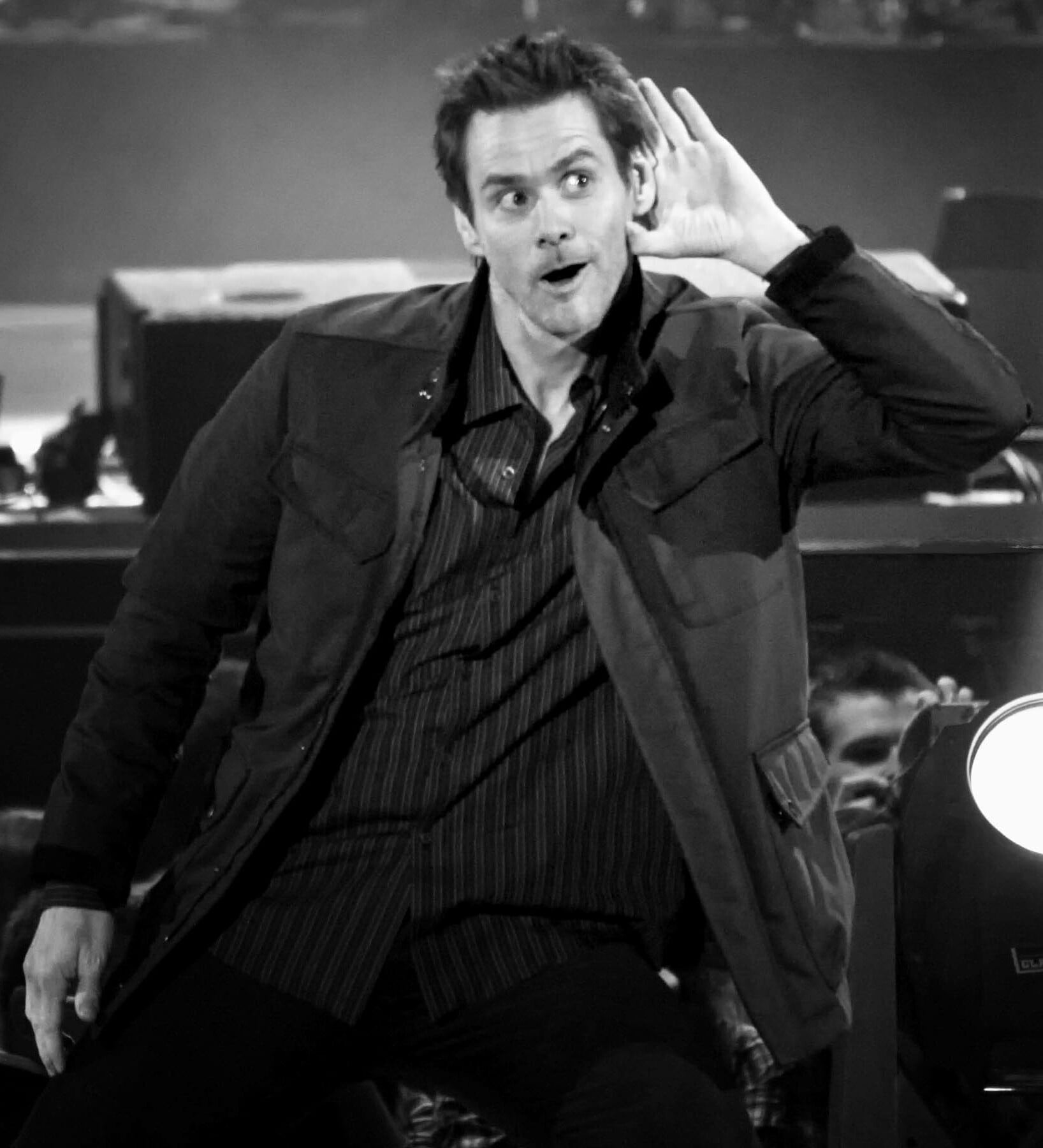 A man in a striped shirt and jacket leans forward on stage, holding a hand to his ear and making an animated, playful facial expression. The image is in black and white.