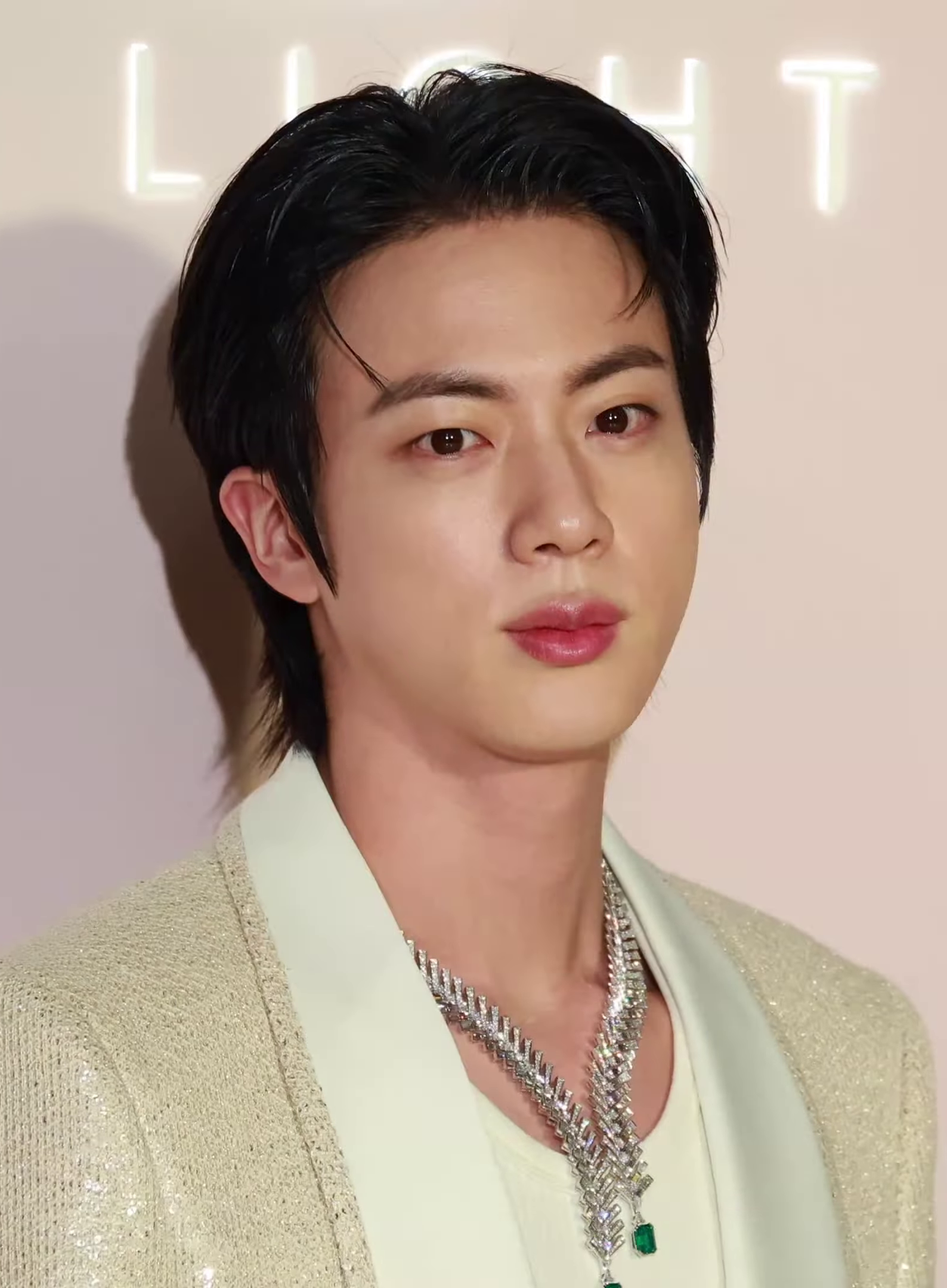 A person with fair skin and dark, straight hair is wearing a light-colored, sparkly jacket and layered silver necklaces, standing in front of a beige background with the word "LIGHT" partially visible.