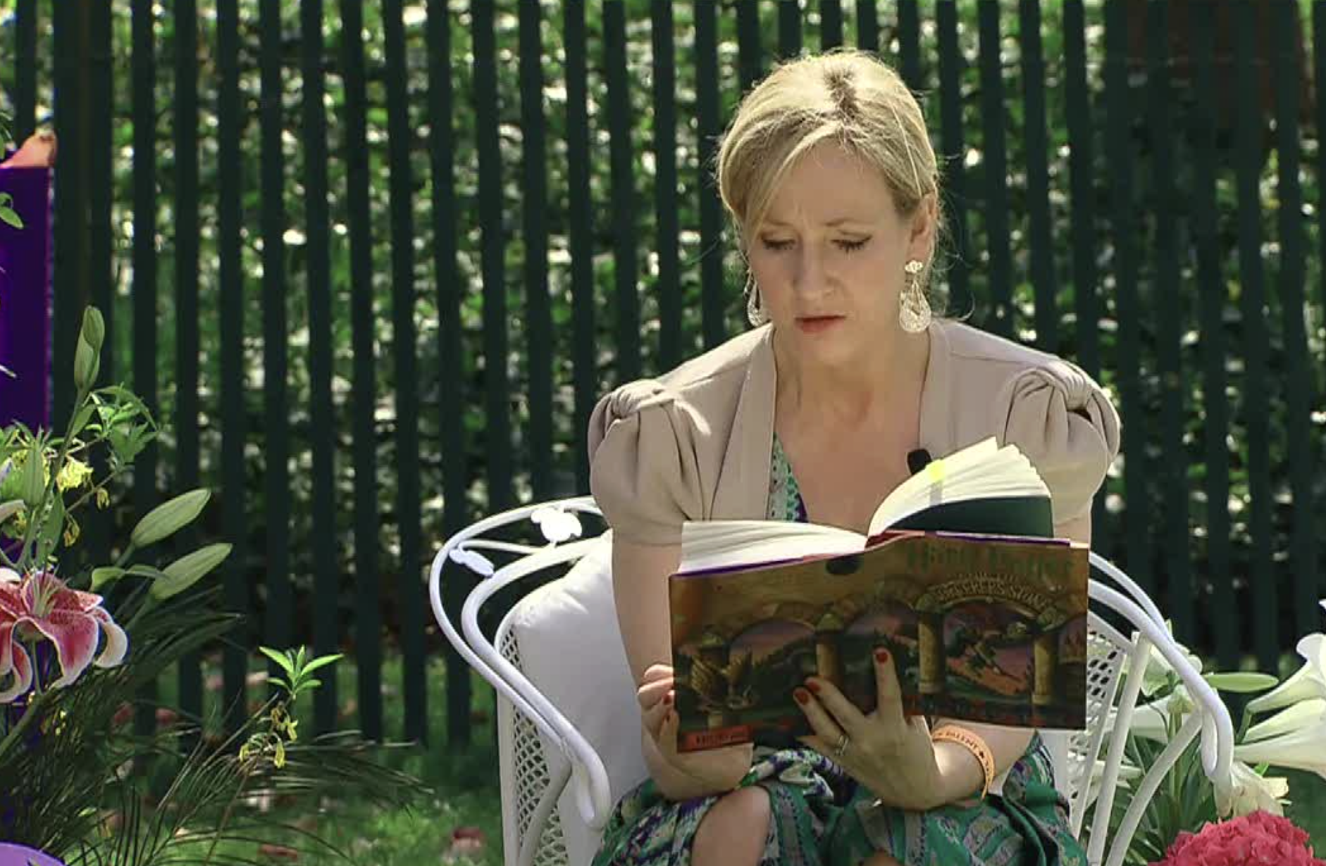 A woman sitting on a white chair in a garden reads a book with a colorful cover. Flowers and greenery surround her, and a green fence is visible in the background.