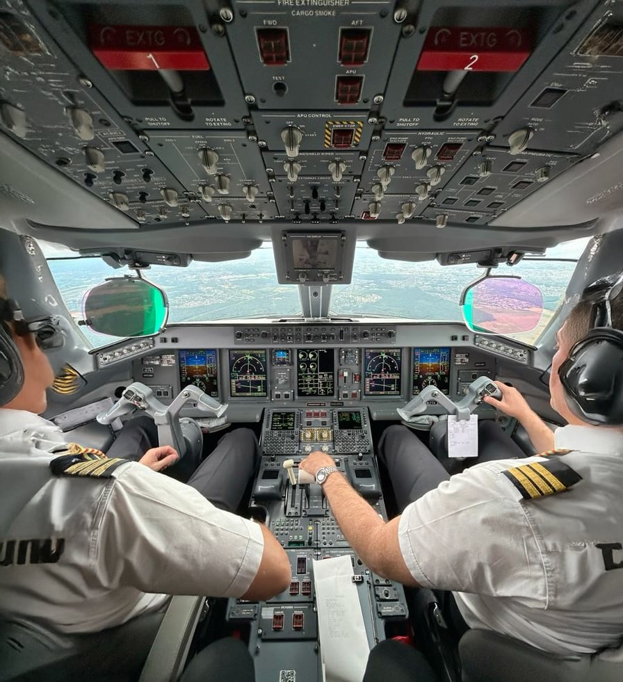 Two pilots in uniform sit in the cockpit of an airplane, operating the controls as the plane approaches a runway for landing. The cockpit is filled with buttons, switches, and digital instrument displays.