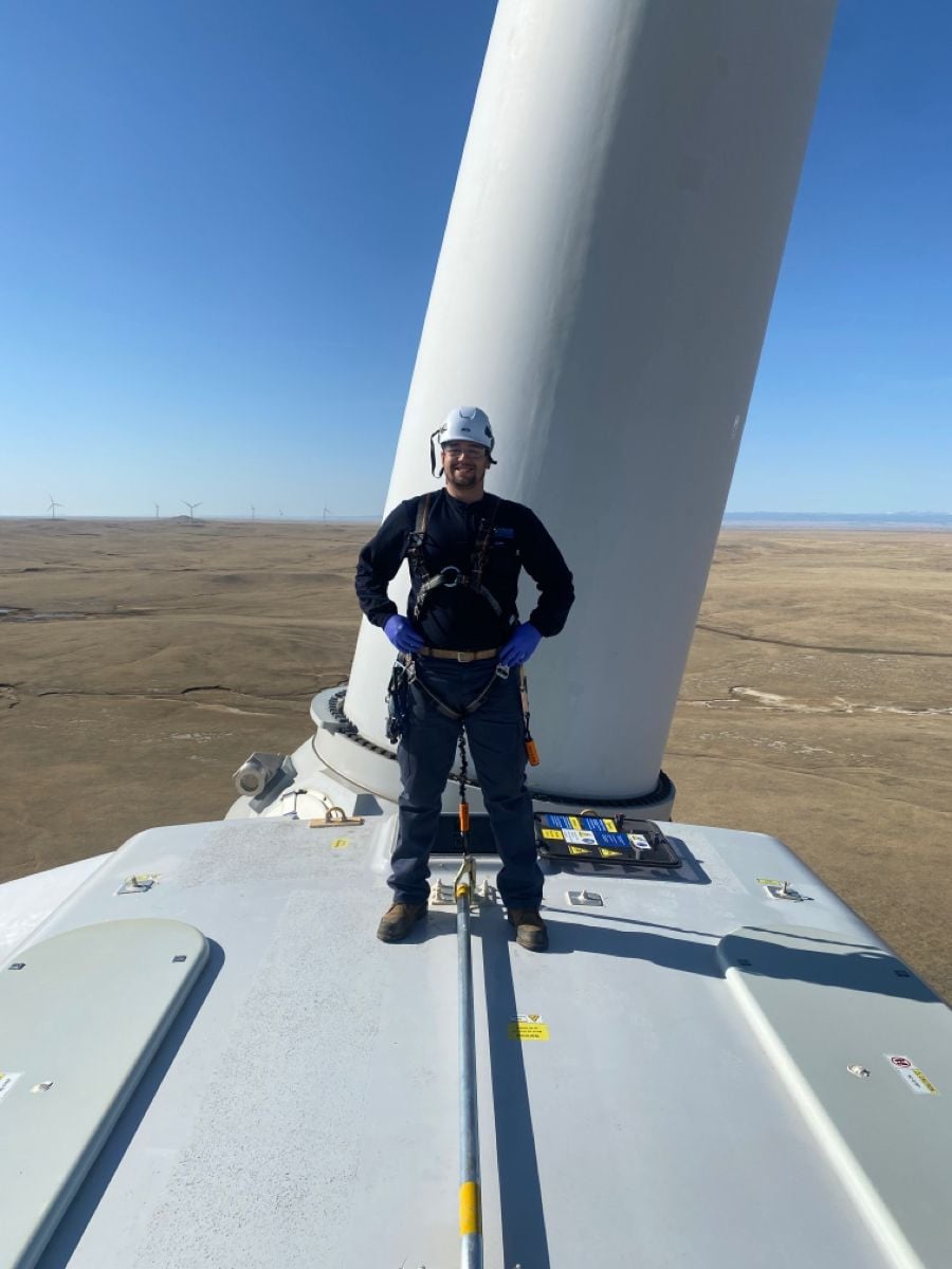 A worker wearing safety gear and a helmet stands on top of a wind turbine with wide open plains and other wind turbines visible in the background under a clear blue sky.
