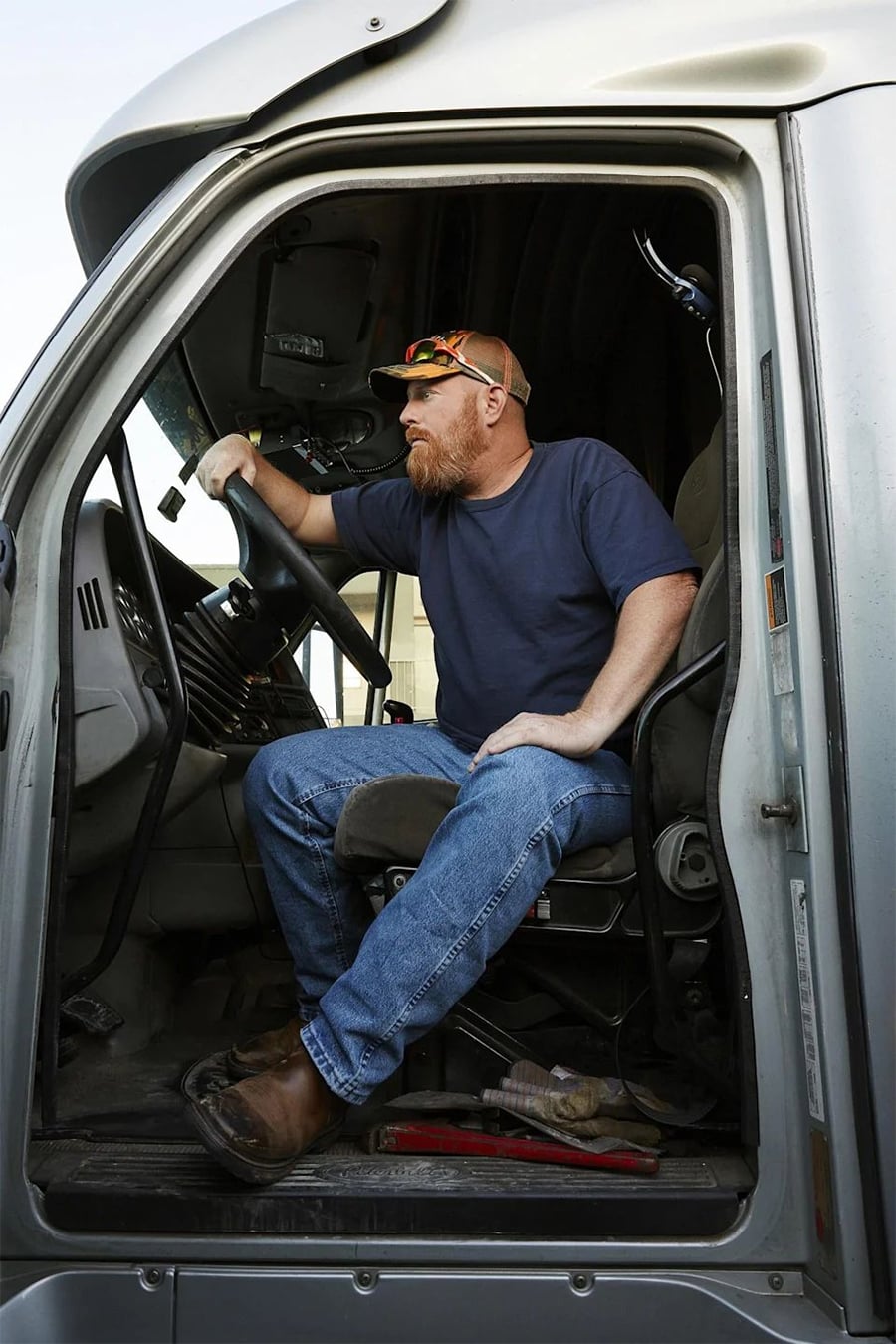 A man with a beard, wearing sunglasses, a cap, a navy t-shirt, and jeans, sits in the driver’s seat of a large truck, looking out the open door with one hand on the steering wheel.