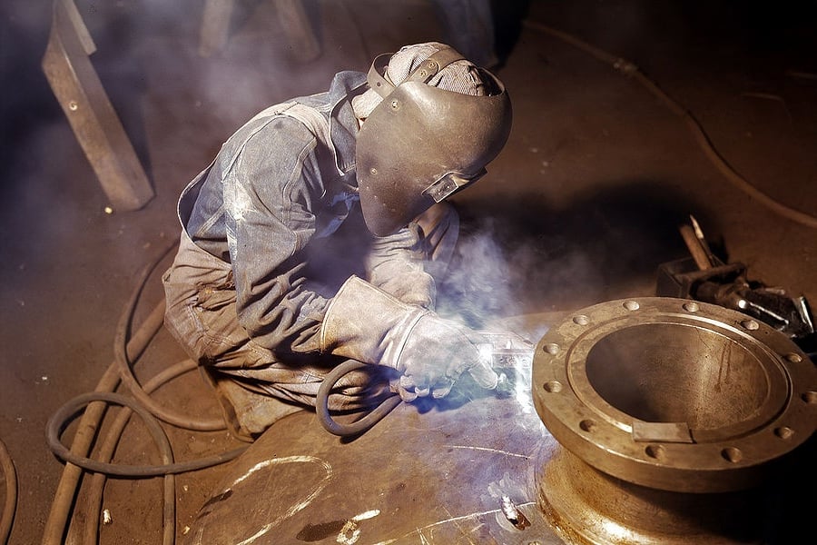 A person wearing protective gear and a welding mask is crouched down, welding a large metal component. Bright sparks and smoke are visible around the welding area, with cables and tools scattered nearby.