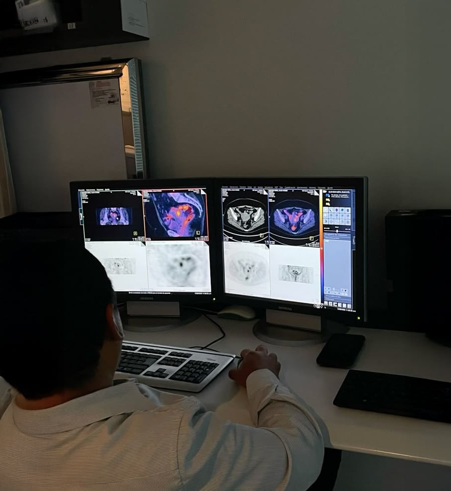 A person sits at a desk analyzing medical scans on two computer monitors, displaying colored and grayscale images, possibly PET or MRI scans. The room is dimly lit, and computer accessories are on the desk.