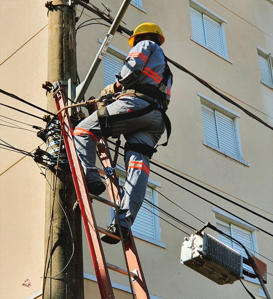 A utility worker wearing a helmet and safety gear stands on a ladder, repairing electrical cables on a utility pole next to a building with closed windows.