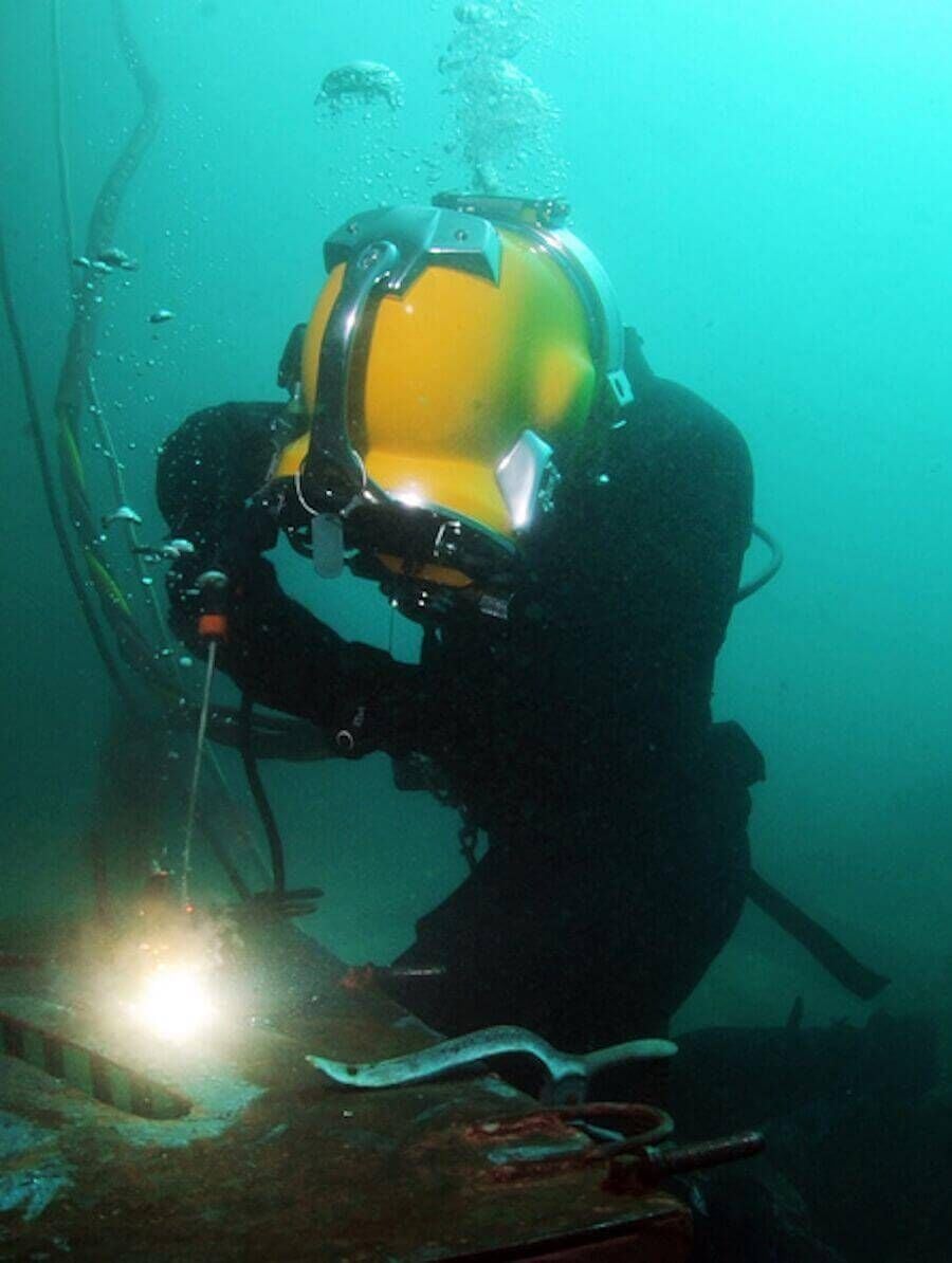 A diver wearing a yellow helmet and protective suit performs underwater welding, with sparks and light visible at the welding point on a submerged metal structure.