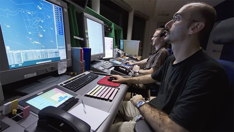 Two air traffic controllers sit at desks with computer monitors displaying flight paths and radar data, using keyboards and headsets to manage air traffic operations in a control room.