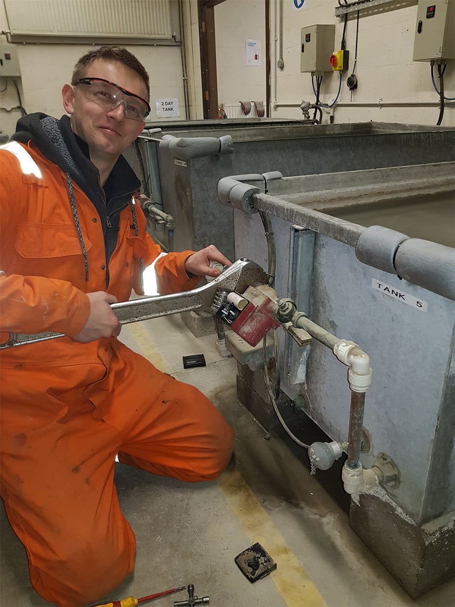 A person wearing orange coveralls and safety glasses kneels beside a large tank, using a large wrench to adjust a valve on the tank’s plumbing in an industrial setting.