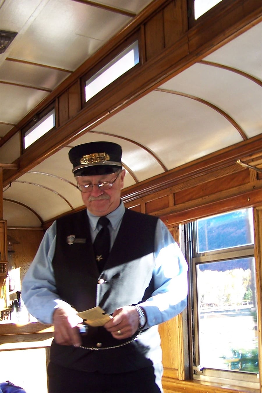 A train conductor in uniform and hat examines a ticket inside a vintage passenger train car with wooden panels and large windows. Sunlight streams through the windows, illuminating the scene.