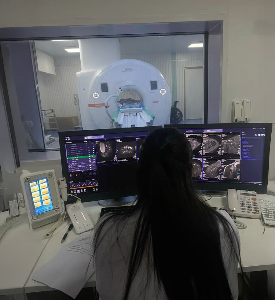 A person with long dark hair sits at a control desk, monitoring multiple screens displaying MRI scan images, with an MRI machine visible through a large window in the background.