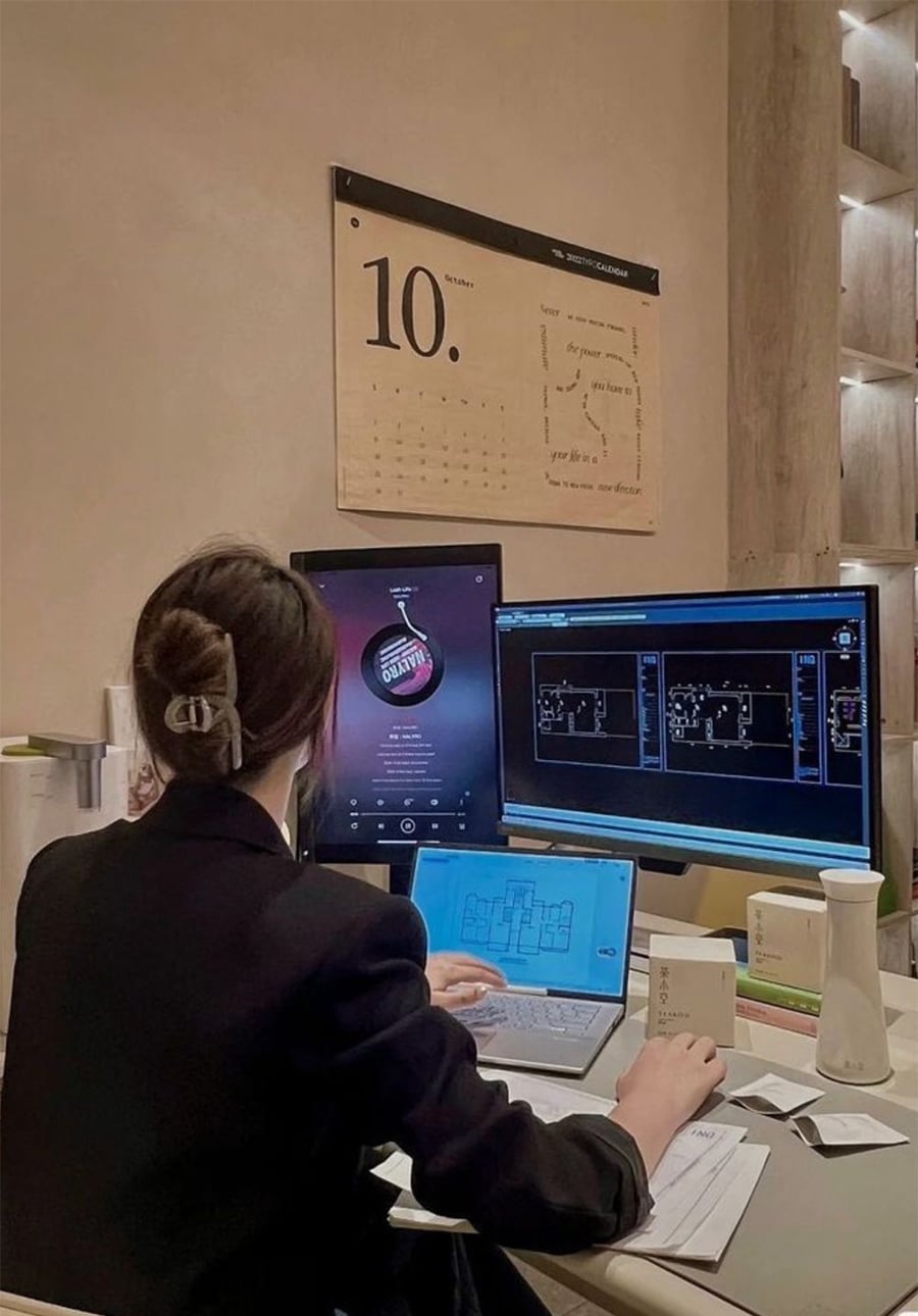 A person with a hair clip sits at a desk using a laptop and two monitors displaying architectural plans. Office supplies and product boxes are on the desk, and a calendar showing October hangs on the wall.