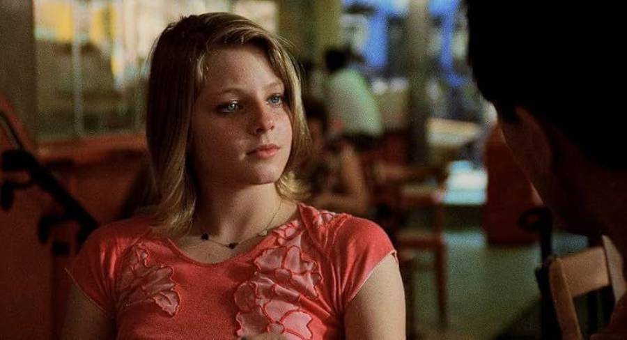 A young woman with blonde hair, wearing a pink shirt with flower designs, sits at a table indoors, looking thoughtfully at someone off-camera. The background is softly blurred, showing chairs and people.