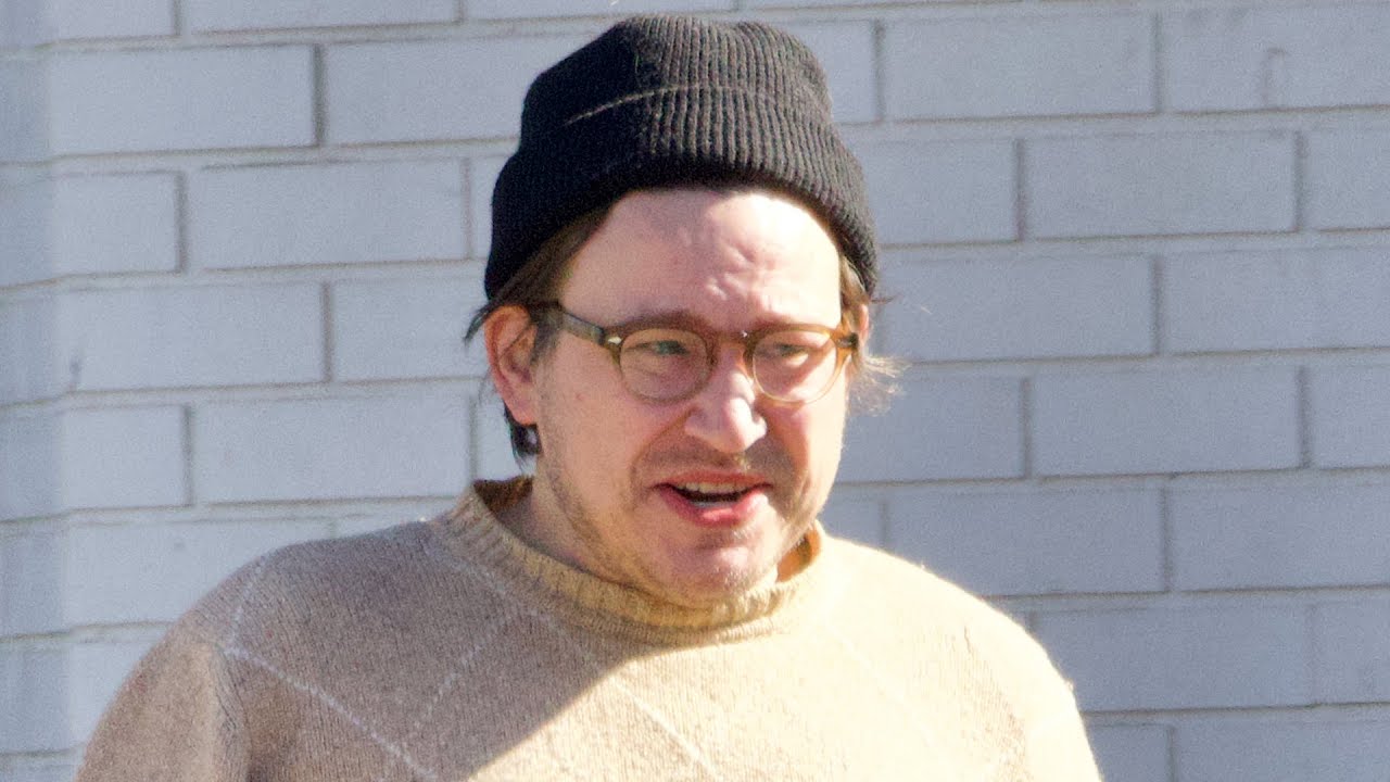 A person wearing glasses, a black beanie, and a beige sweater stands in front of a light-colored brick wall.