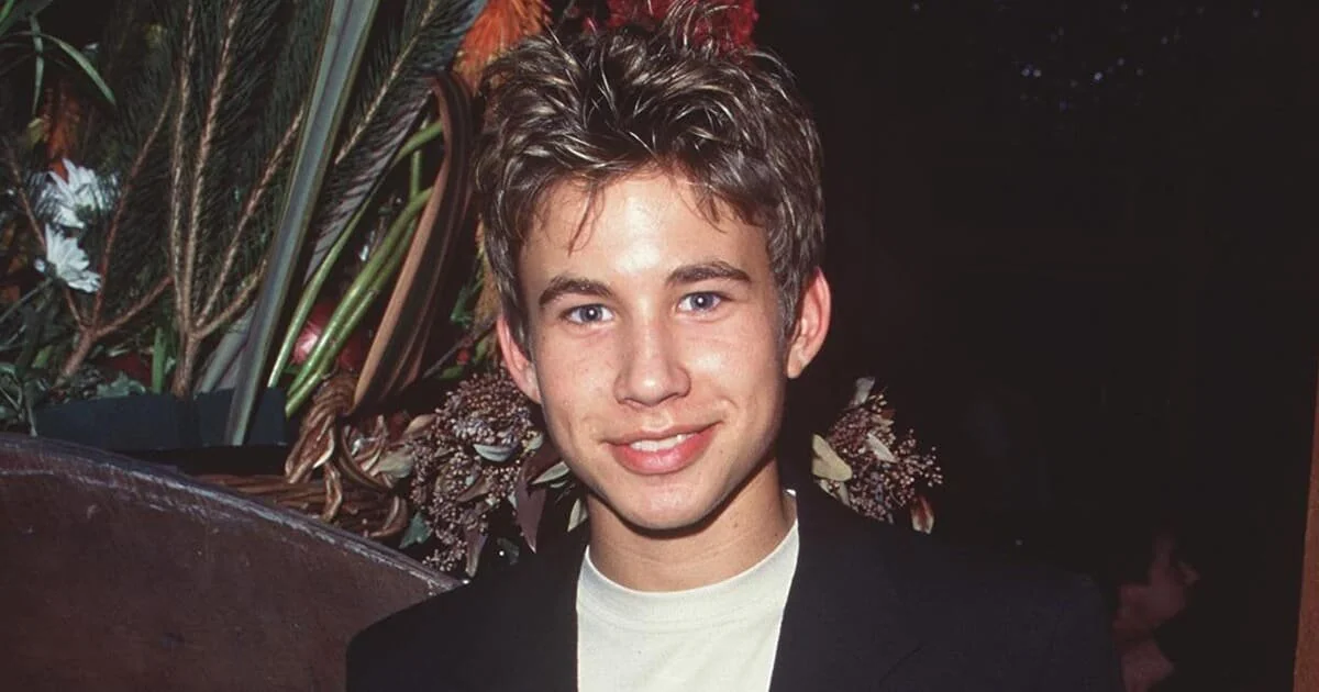 A young person with short, spiky brown hair smiles at the camera, wearing a black jacket over a light-colored shirt. The background features green plants and flowers.