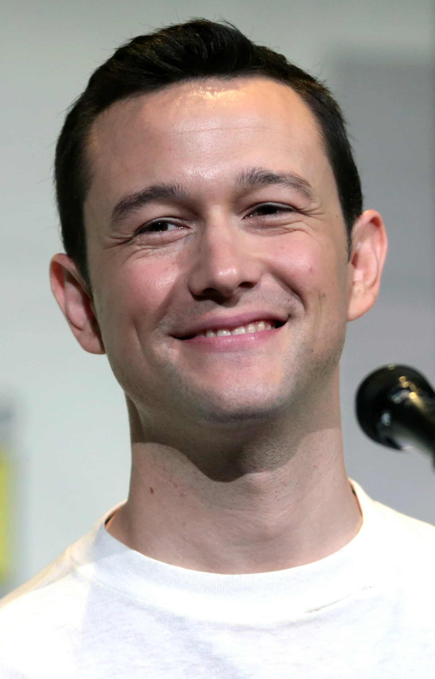 A man with short dark hair smiles while standing in front of a microphone. He is wearing a white shirt and is photographed against a blurred, light-colored background.