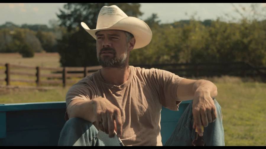 A man with a beard wearing a white cowboy hat and tan t-shirt sits outdoors on the back of a blue truck, holding sunglasses, with a wooden fence and trees in the background.