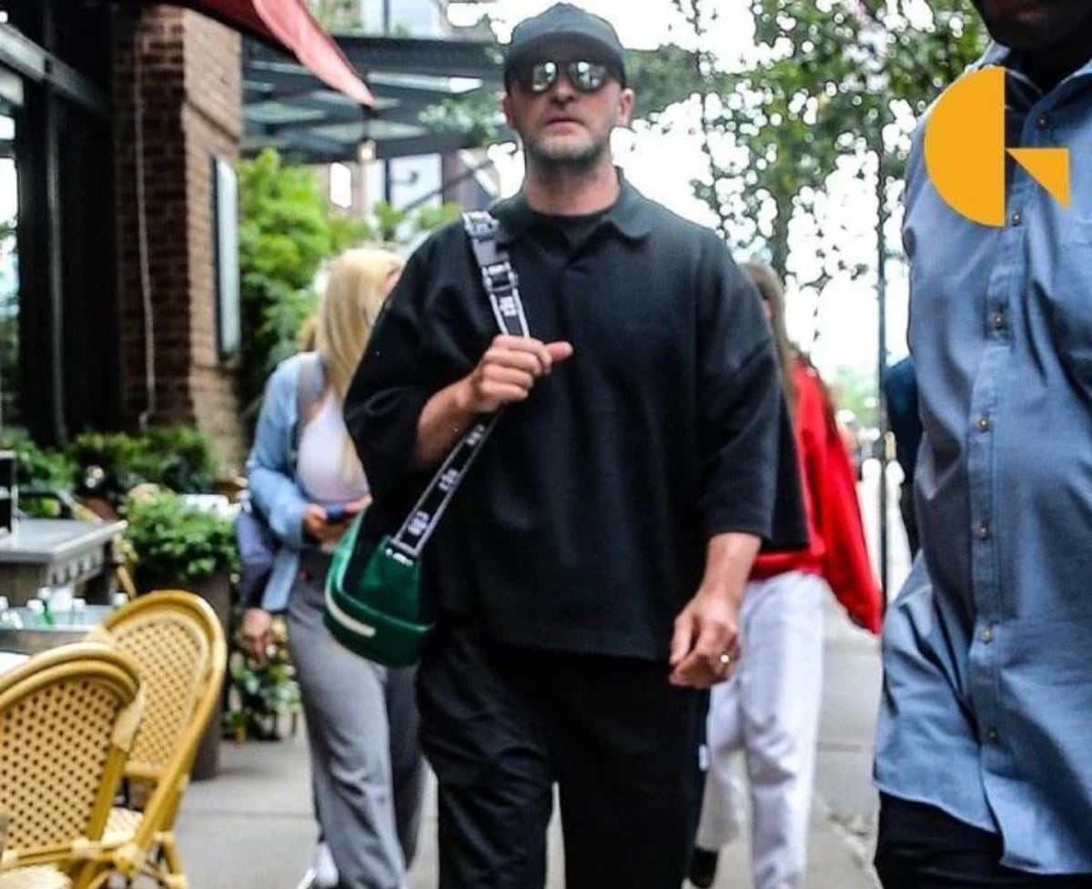 A man wearing a black cap, sunglasses, and black clothing walks on a city sidewalk, carrying a green shoulder bag. There are people and outdoor café chairs in the background.