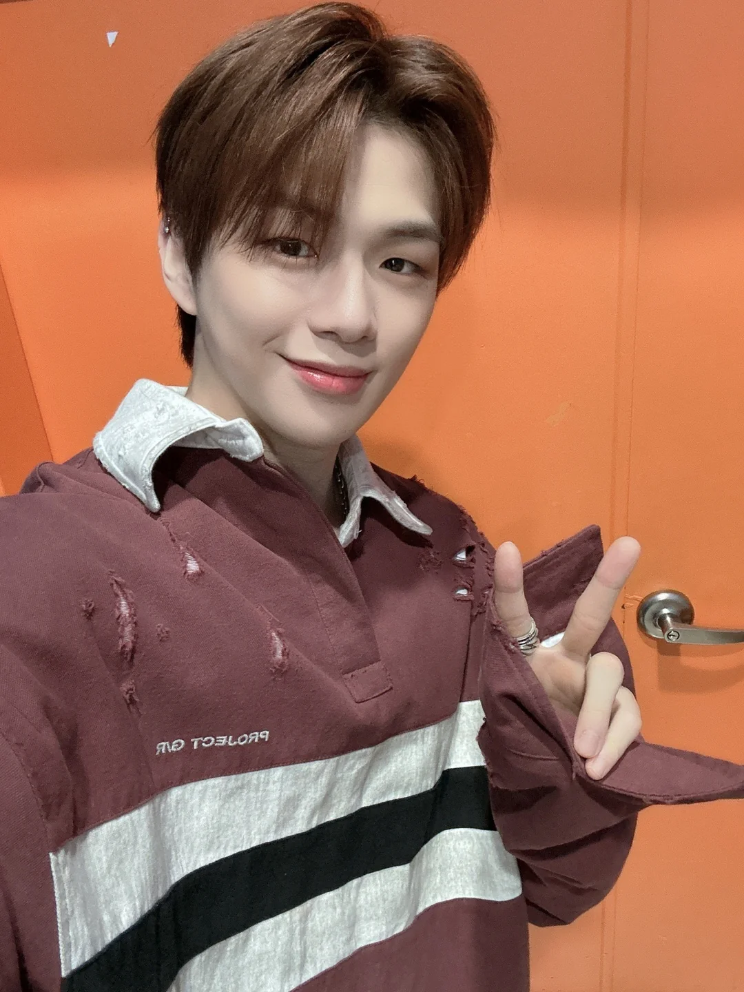 A young person with light brown hair wearing a maroon, white, and black striped shirt poses for a selfie in front of an orange door, smiling and making a peace sign with their right hand.