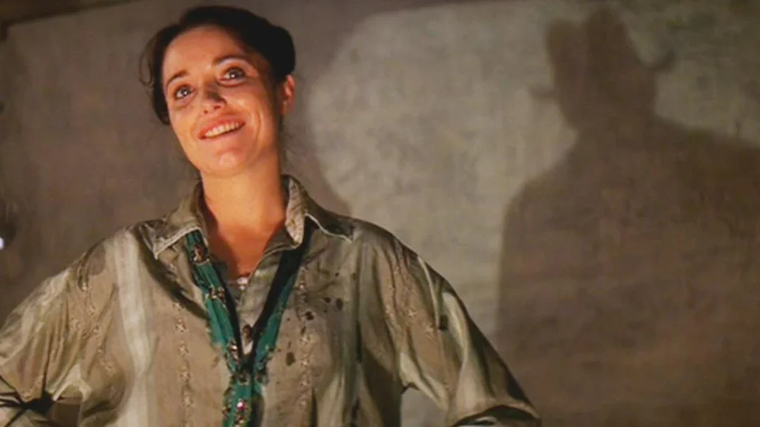 A woman wearing a dirty, loose button-up shirt and green necklace stands smiling in dim lighting, while a large, shadowy figure in a hat is cast on the wall behind her.
