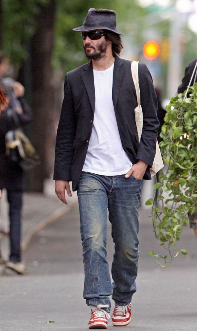 A man wearing a black fedora, sunglasses, black blazer, white t-shirt, blue jeans, and red sneakers walks on a city sidewalk with one hand in his pocket and a tote bag over his shoulder.