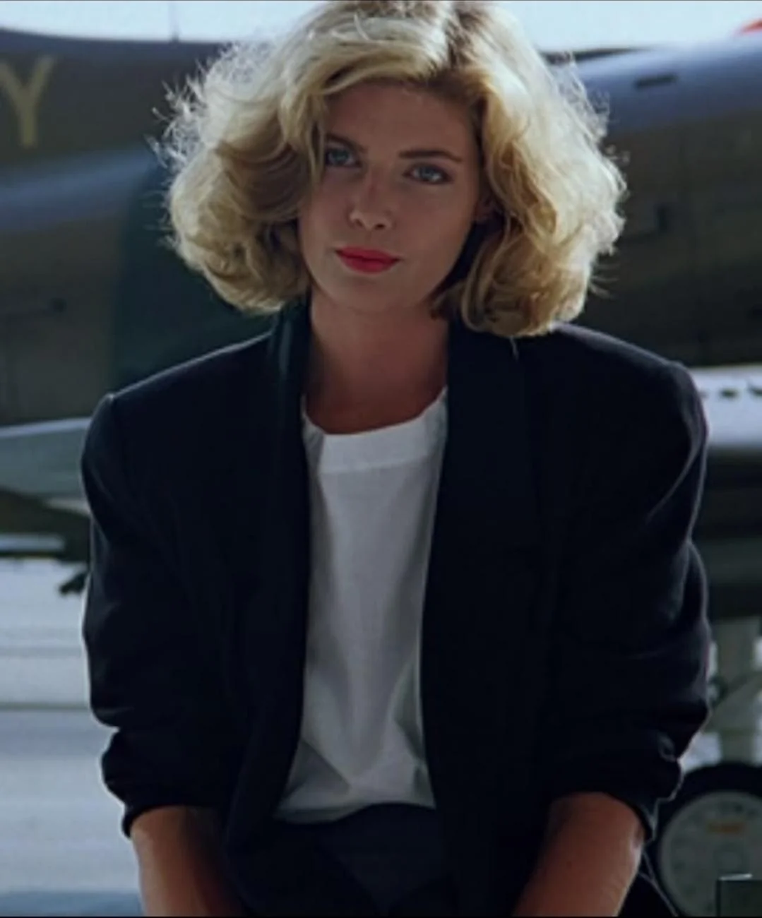 A woman with blonde, wavy hair wearing a black blazer and white shirt stands in front of an aircraft, looking at the camera with a neutral expression.