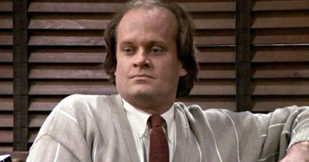 A man with thinning hair, wearing a light-colored cardigan and a red tie, sits in front of wooden blinds, looking slightly to the side with a neutral expression.