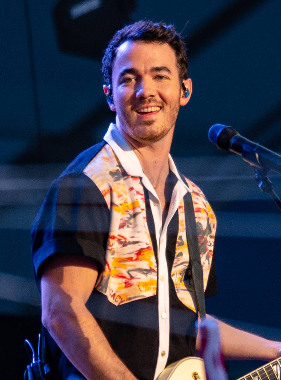 A man with short dark hair, wearing a colorful short-sleeved shirt and holding a guitar, stands smiling in front of a microphone on stage.