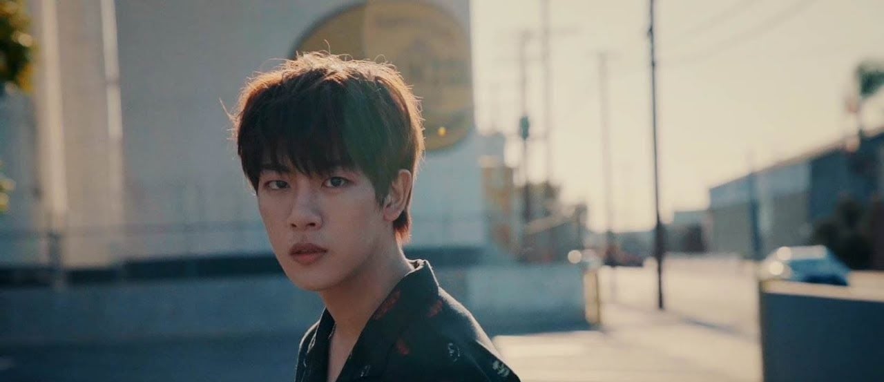 A young person with short, dark hair stands outdoors in soft sunlight, looking toward the camera. The background shows a blurred street scene with buildings and power lines.