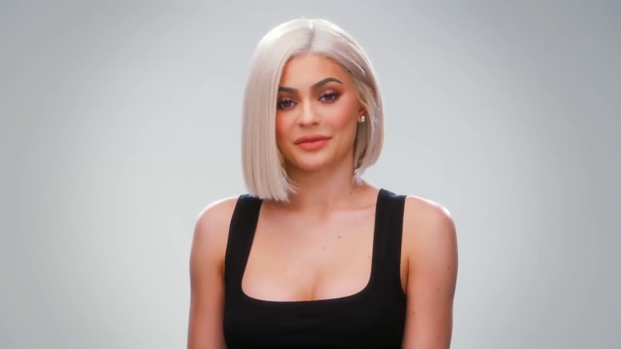 A woman with straight platinum blonde hair and a blunt bob is wearing a black sleeveless top and subtle makeup. She is looking forward with a neutral expression against a plain light grey background.