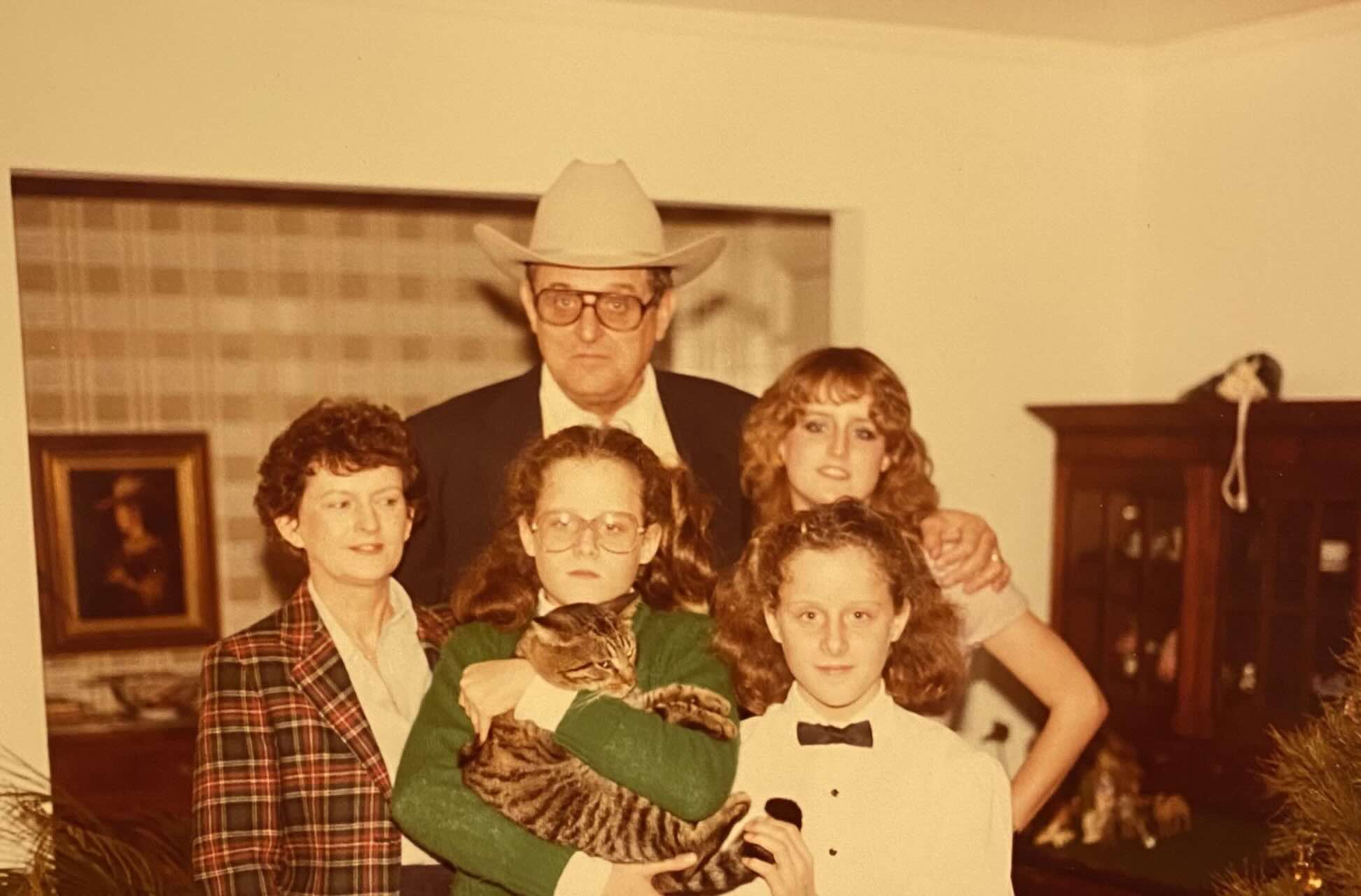 A vintage family photo shows two adults and three children posing indoors. One child holds a tabby cat. The man wears glasses and a cowboy hat; the others wear plaid, green, and white outfits. They stand in a warmly-lit room.