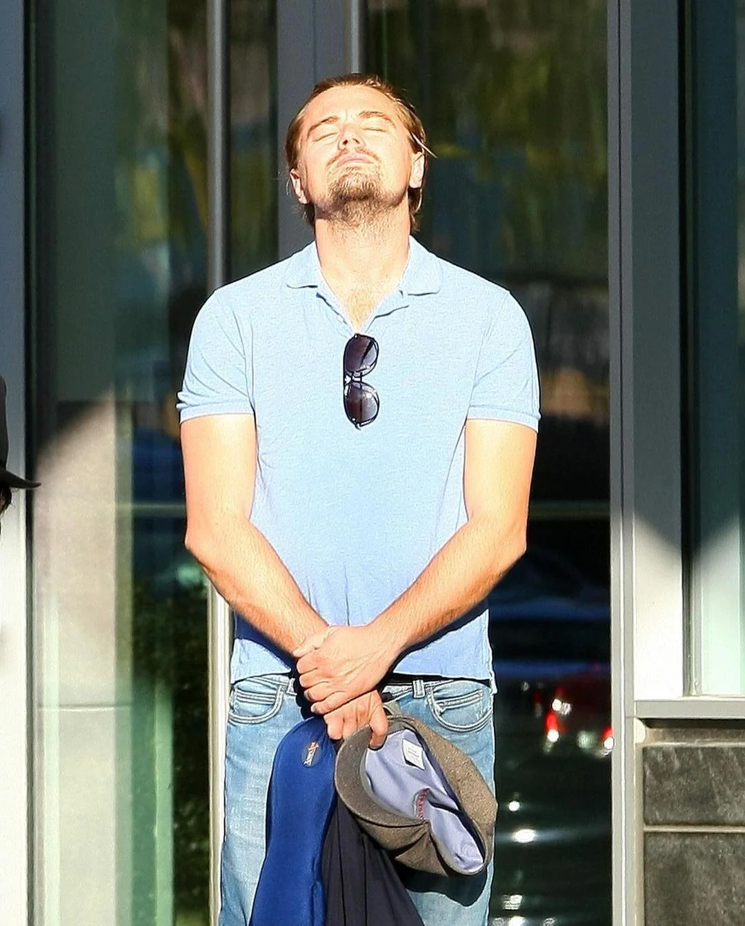 A man wearing a light blue polo shirt and jeans stands outside a building, eyes closed, facing the sun with a relaxed expression. He holds a jacket and hat in his hands, and sunglasses hang from his shirt.