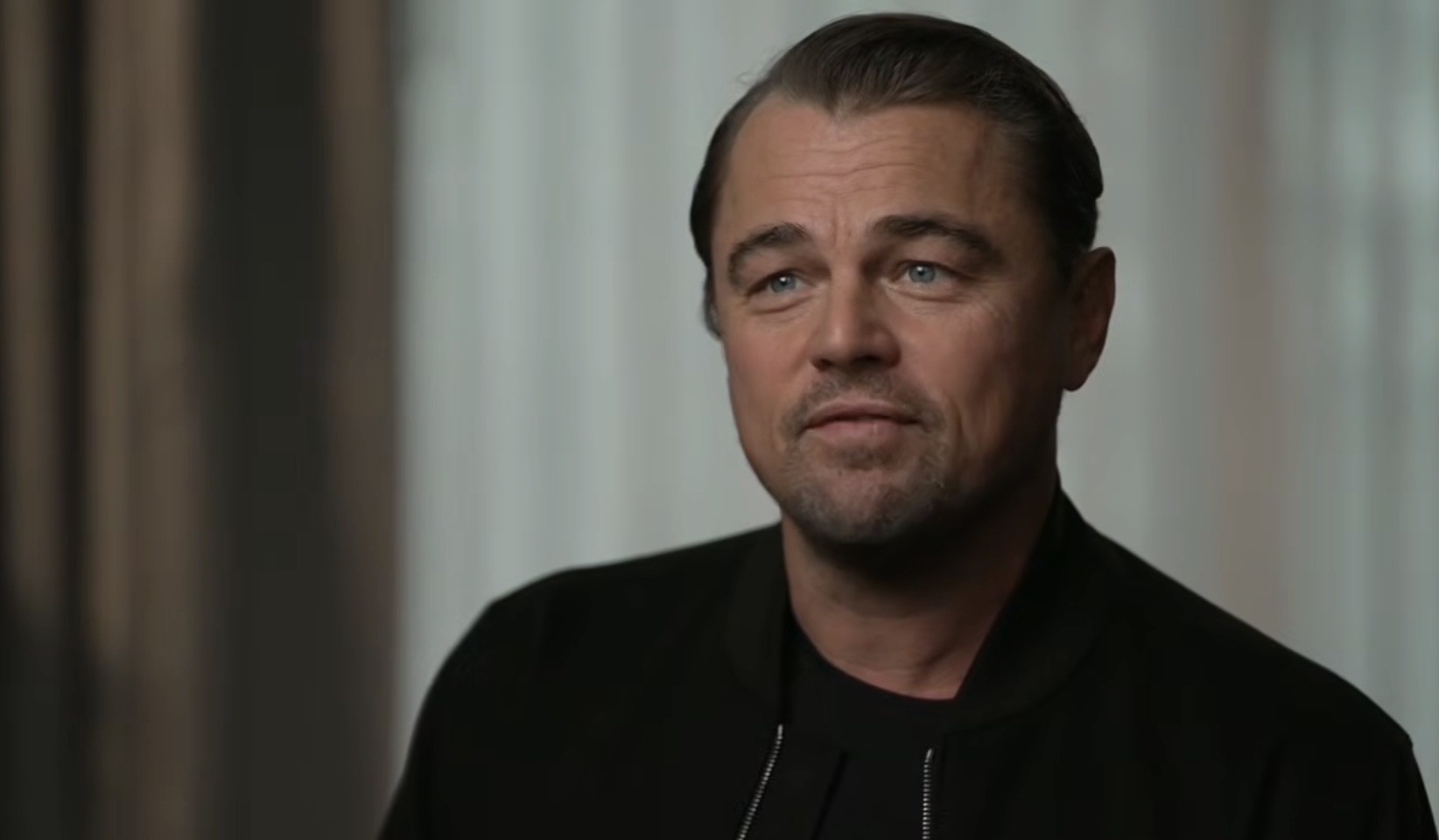 A man with slicked-back hair and a trimmed beard, wearing a black jacket over a black shirt, sits indoors against a softly blurred background with curtains and natural light.