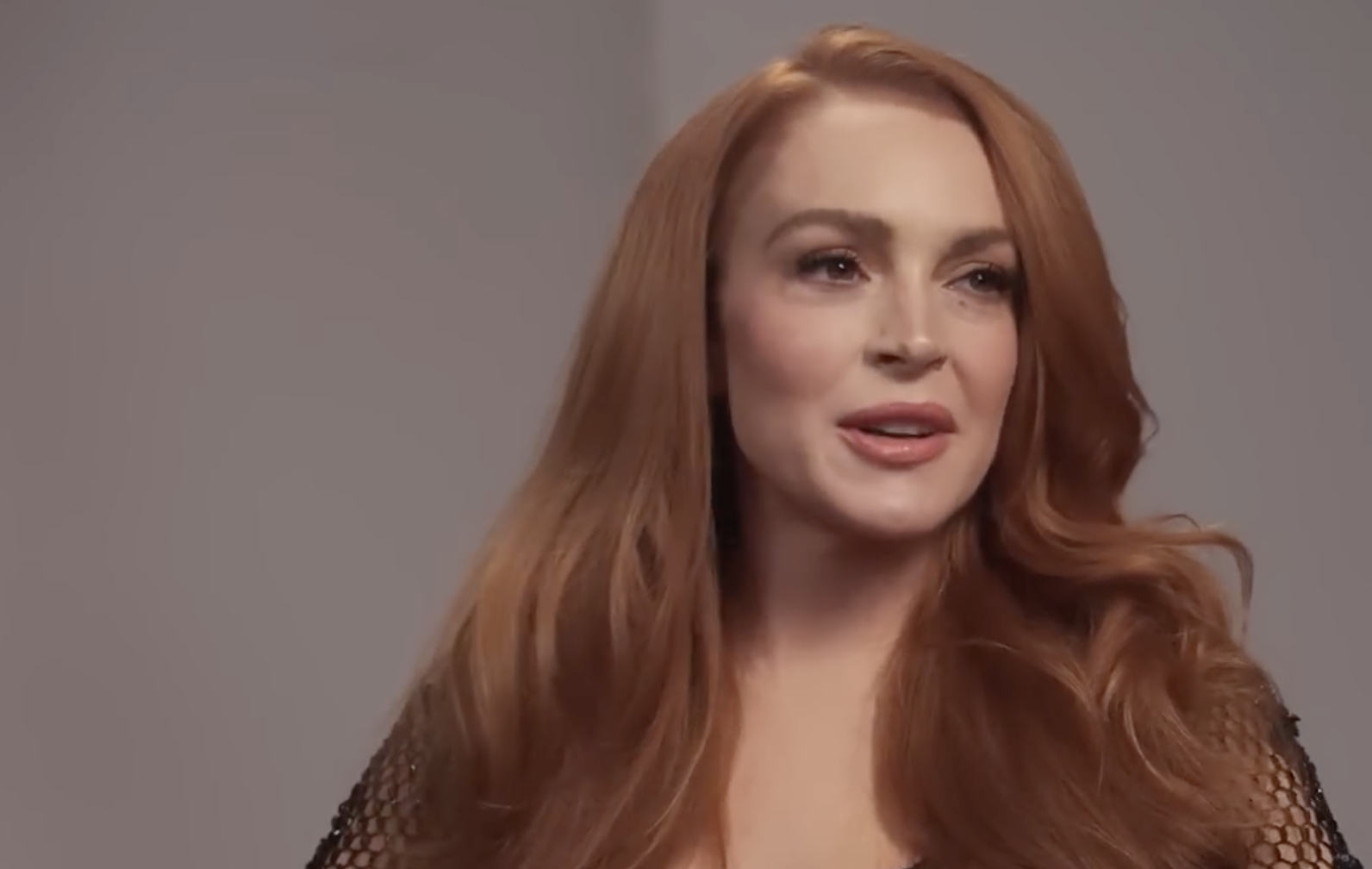 A woman with long, wavy red hair is looking slightly to the side and smiling softly. She is wearing a black, textured top and is in front of a plain, light-colored background.