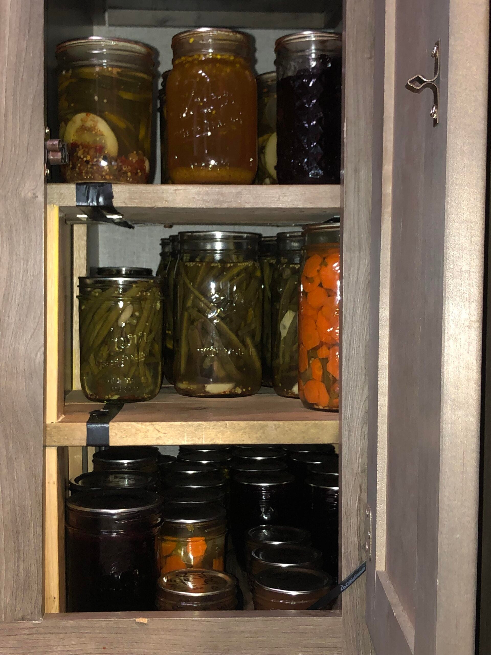 A pantry cabinet with three shelves filled with various jars of home-canned foods, including green beans, carrots, pickles, jam, and preserves. The jars are of different sizes and tightly packed together.