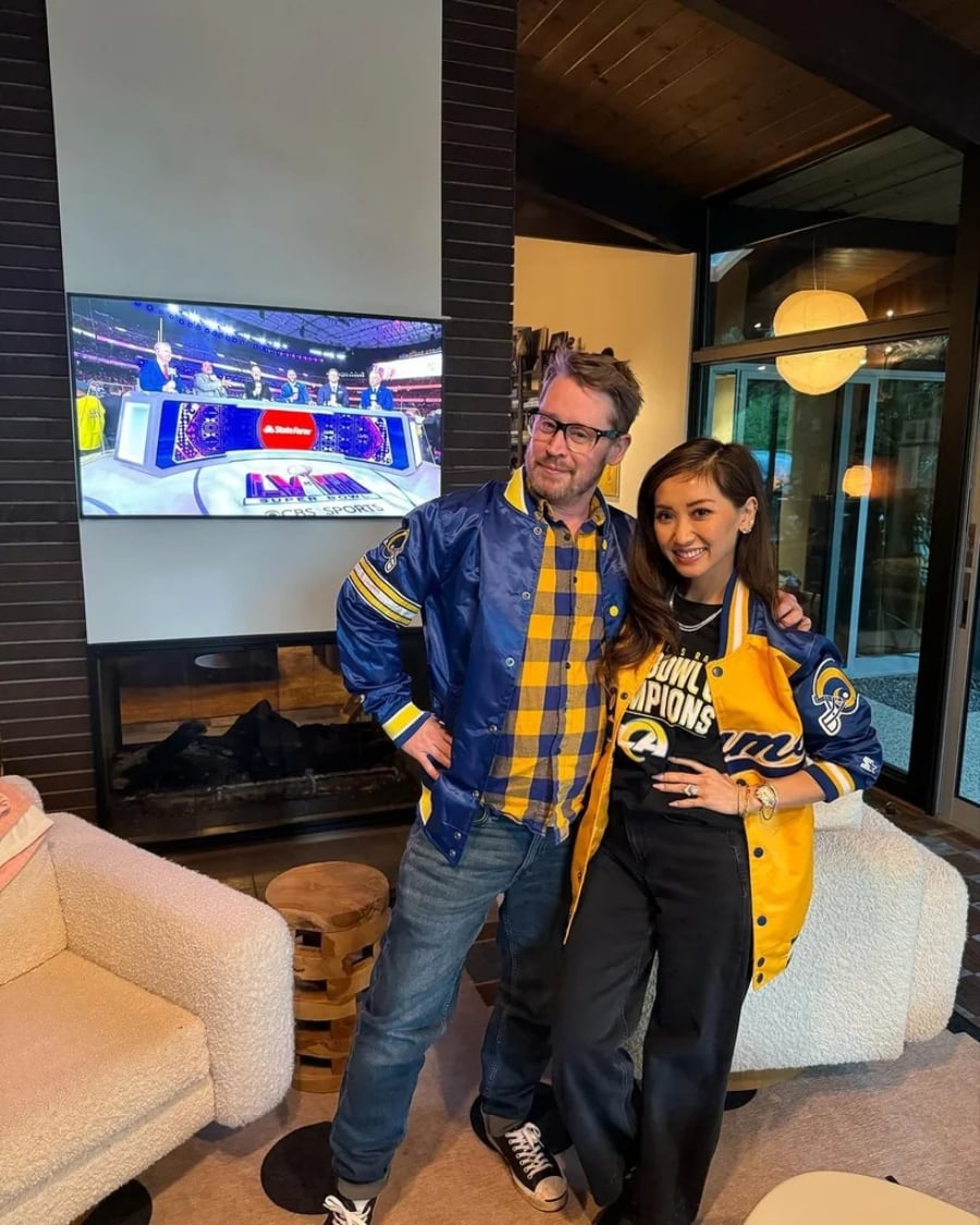 Two people smile and pose indoors wearing Los Angeles Rams jackets; a football game with a halftime show is visible on the TV behind them. The room has cozy seating, a fireplace, and large windows.