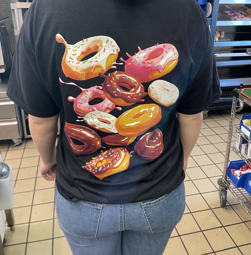 A person wearing a black T-shirt with colorful, realistic illustrations of assorted donuts on the back, standing indoors on a tiled floor. Shelves with supplies and equipment are visible in the background.