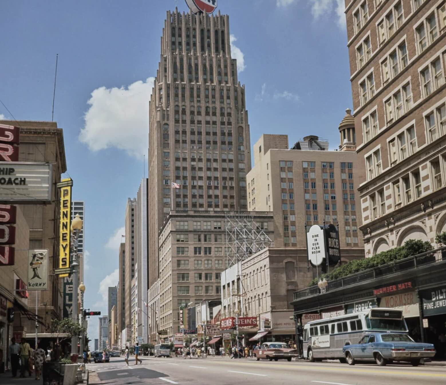 A busy city street with tall buildings, vintage cars, buses, and storefront signs, including Penney’s. The scene suggests a mid-20th century urban setting under a clear, sunny sky.