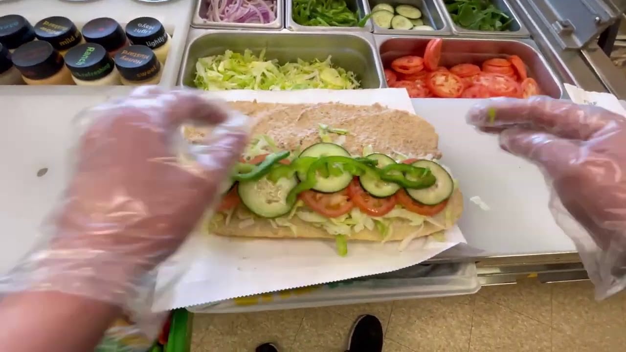 A person wearing gloves assembles a sub sandwich with lettuce, tomato, cucumber, and green bell pepper at a sandwich shop, with various fresh ingredients and sauces visible in metal containers.