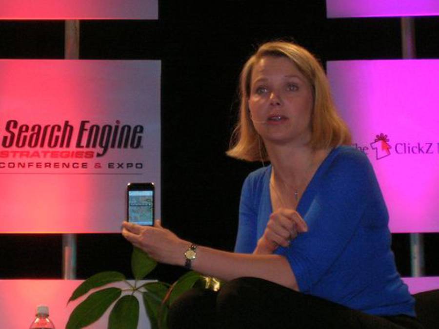 A woman in a blue top sits in front of "Search Engine Strategies Conference & Expo" signage, holding up a smartphone and speaking, with colorful lights in the background.