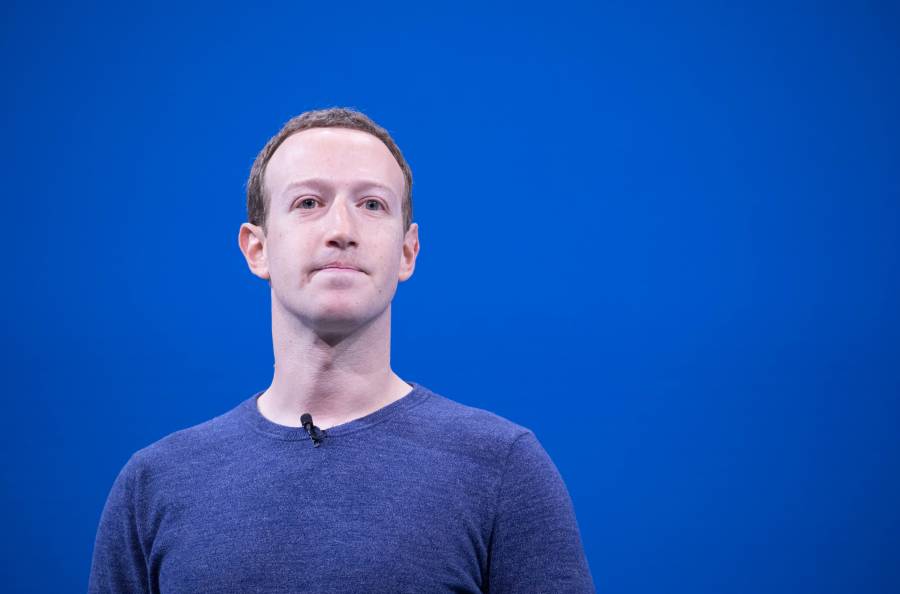 A man with short brown hair wearing a dark blue sweater stands in front of a solid blue background, looking slightly to the side. He has a microphone attached to his shirt.