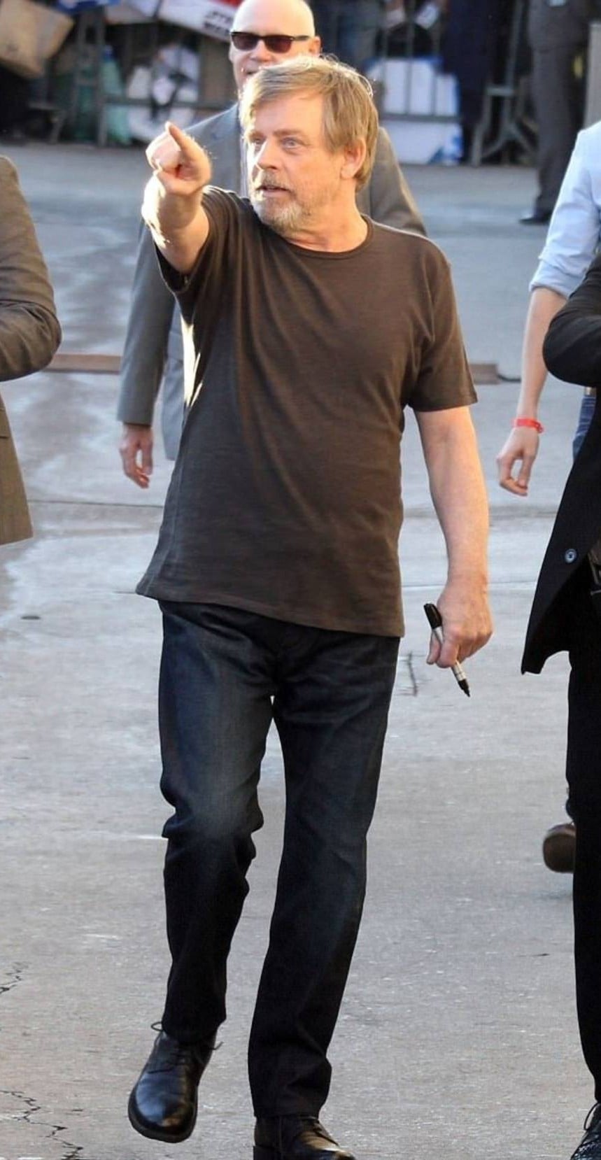 A man with light hair and a beard wearing a dark t-shirt, jeans, and black boots is walking outside while pointing forward. He holds glasses in one hand and people are visible in the background.