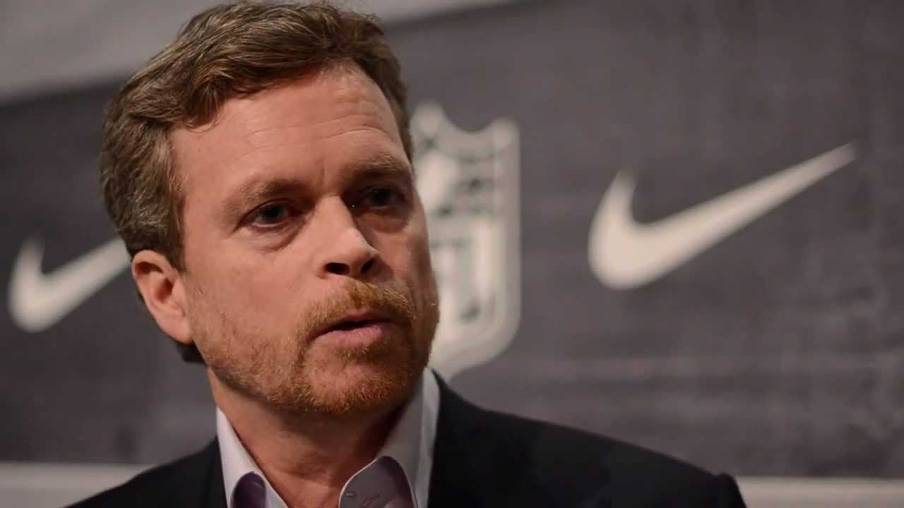 A man with light brown hair and a beard, wearing a suit jacket and light shirt, is speaking in front of a backdrop featuring the NFL logo and Nike swoosh symbols.