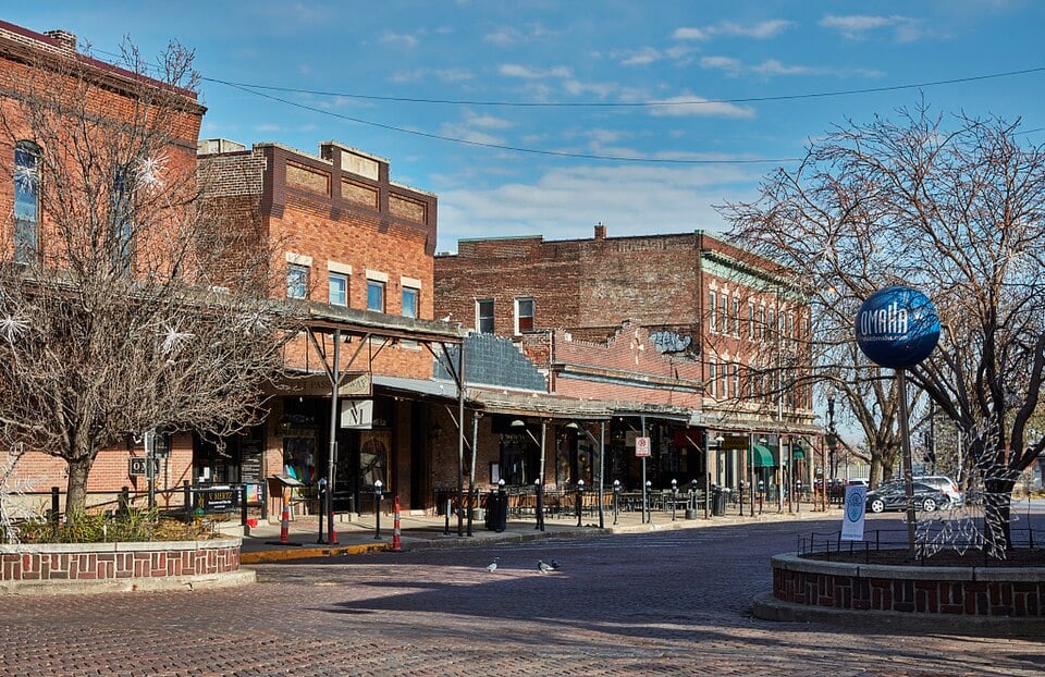 A brick-paved street lined with historic red-brick buildings, bare trees, outdoor seating, and a blue sphere sculpture on a pedestal, all under a partly cloudy sky.